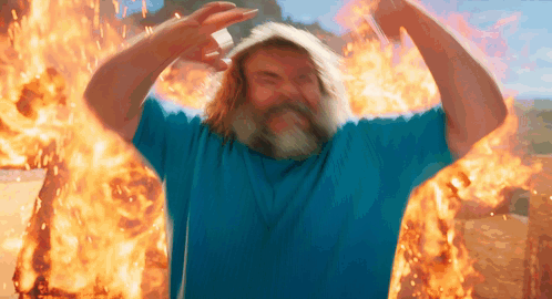 A Man With A Beard And A Blue Shirt Is Standing In Front Of A Fire GIF