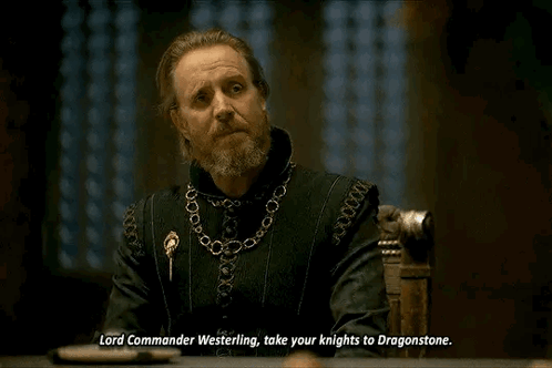 a man with a beard and a chain around his neck is sitting at a table and talking to someone .
