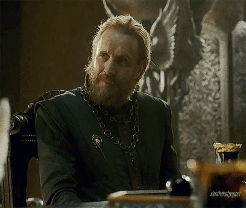 a man with a beard and a chain around his neck is sitting at a table in a chair .