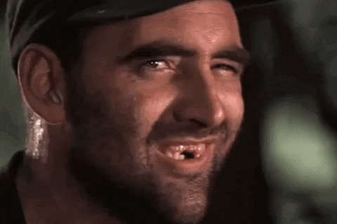 A Man With A Beard And A Hat Is Smiling With A Hole In His Mouth . GIF