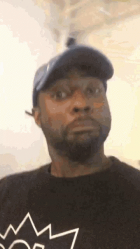 A Man With A Beard And A Hat Is Wearing A Black Shirt With A Crown On It . GIF