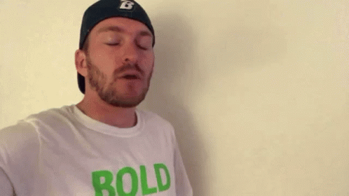 A Man With A Beard And A Hat Is Wearing A White T-Shirt That Says `` Bold '' . GIF