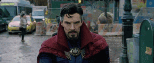 A Man With A Beard And A Red Cape Is Standing In A Street . GIF