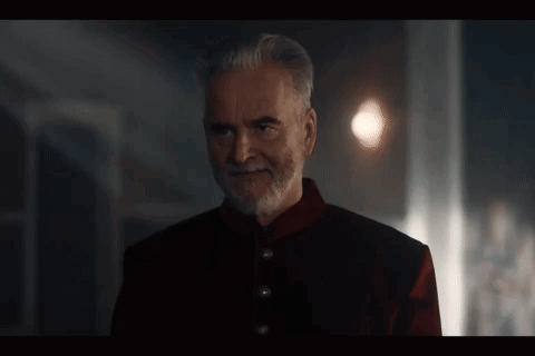 A Man With A Beard And A Red Jacket Is Standing In A Dark Room And Smiling . GIF