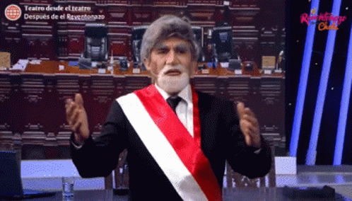 A Man With A Beard And A Red White And Black Sash Is Standing In Front Of A Sign That Says Teatro Desde El Teatro GIF