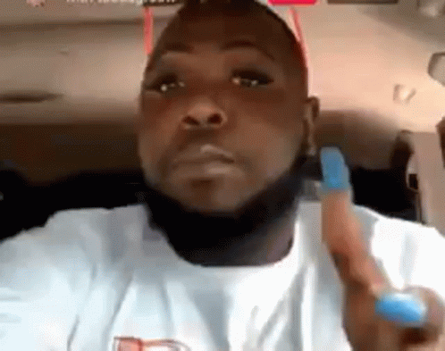 A Man With A Beard And A White Shirt Is Sitting In A Car Holding A Blue Object . GIF