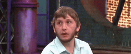 A Man With A Beard And A White Shirt Is Sitting In Front Of A Purple Wall . GIF