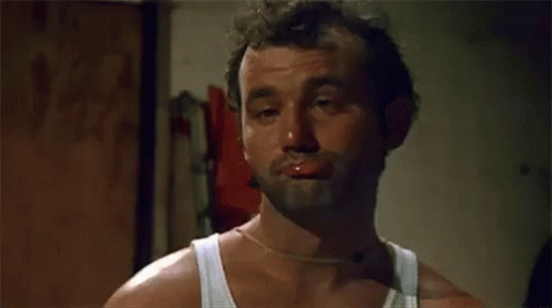 A Man With A Beard And A White Tank Top Is Making A Funny Face With His Mouth Open . GIF