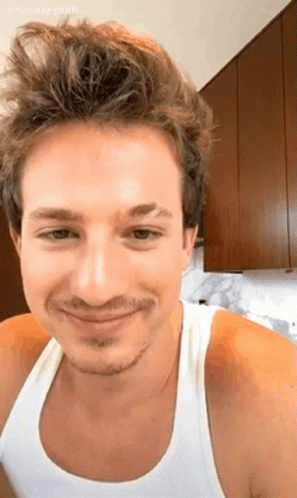 A Man With A Beard And A White Tank Top Is Smiling In A Kitchen . GIF