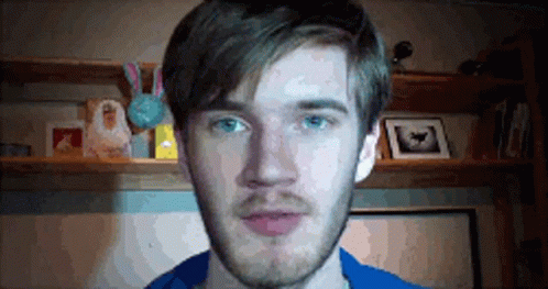 A Man With A Beard And Blue Eyes Is Standing In Front Of A Shelf With Pictures On It . GIF