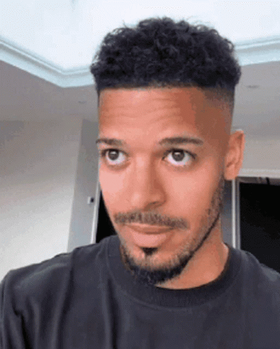 A Man With A Beard And Curly Hair Is Wearing A Black Shirt And Looking At The Camera . GIF