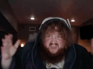 A Man With A Beard And Curly Hair Is Wearing A Hooded Jacket And Making A Funny Face . GIF