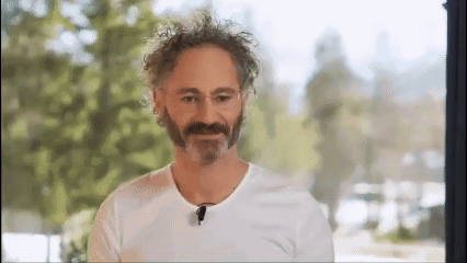 A Man With A Beard And Curly Hair Is Wearing A White T-Shirt And A Microphone . GIF