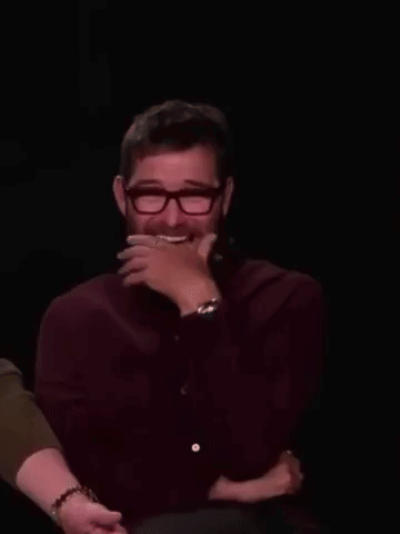 A Man With A Beard And Glasses Is Laughing With His Hand On His Face . GIF