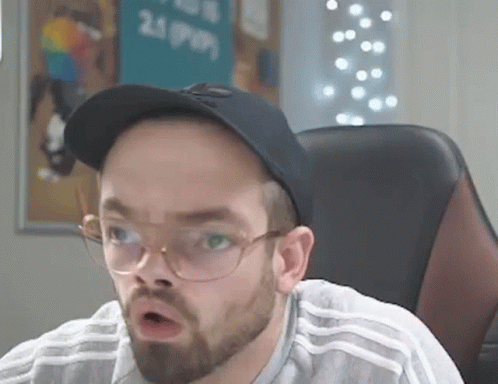 A Man With A Beard And Glasses Is Sitting In A Chair Wearing A Hat And Glasses . GIF