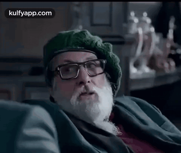 A Man With A Beard And Glasses Is Sitting On A Couch Wearing A Green Hat . GIF