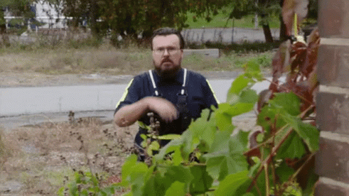 A Man With A Beard And Glasses Is Standing In Front Of A Plant . GIF
