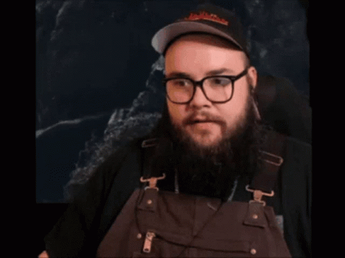 A Man With A Beard And Glasses Is Wearing Overalls And A Hat GIF