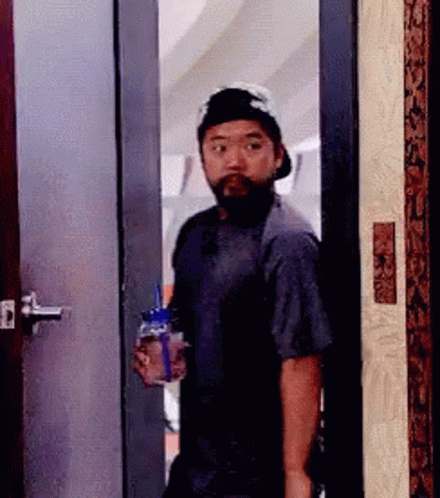 A Man With A Beard And Hat Is Standing In A Doorway Holding A Bottle Of Water . GIF