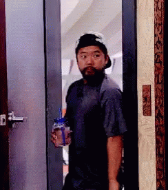 A Man With A Beard And Hat Is Standing In A Doorway Holding A Bottle Of Water . GIF