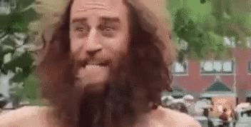 A Man With A Beard And Long Hair Is Standing In Front Of A Building And Making A Funny Face . GIF