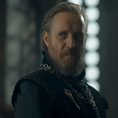 a man with a beard and long hair is wearing a black vest and a gold chain around his neck .