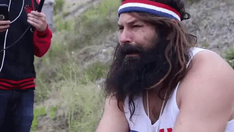 A Man With A Beard And Long Hair Is Wearing A Headband And A White Tank Top . GIF