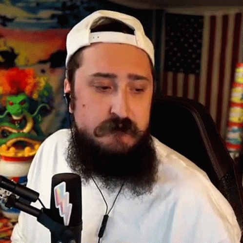 A Man With A Beard And Mustache Is Sitting In Front Of A Microphone Wearing Headphones And A White Hat . GIF