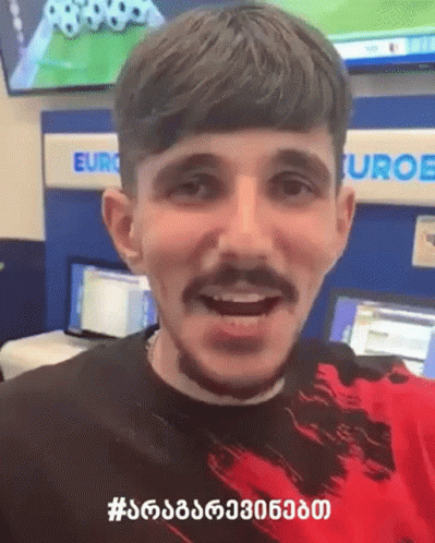 A Man With A Beard And Mustache Is Smiling In Front Of A Sign That Says Euro GIF
