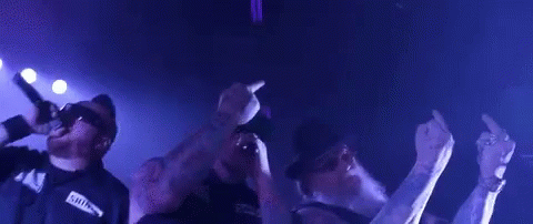 A Man With A Beard And Sunglasses Is Singing Into A Microphone While Another Man Stands Behind Him . GIF