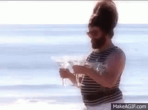 A Man With A Beard And Sunglasses Is Standing On A Beach Holding A Glass Of Water . GIF