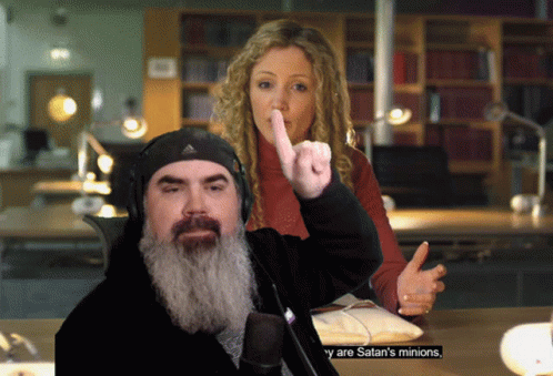 A Man With A Beard Giving The Middle Finger While A Woman Holds Her Finger To Her Mouth GIF