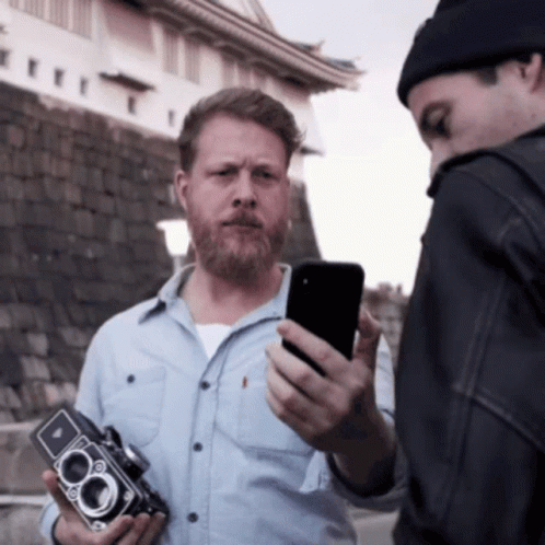 A Man With A Beard Holds A Camera While Another Man Looks At His Phone GIF