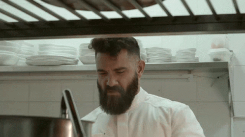 A Man With A Beard In A Kitchen With Plates On The Shelf Behind Him GIF