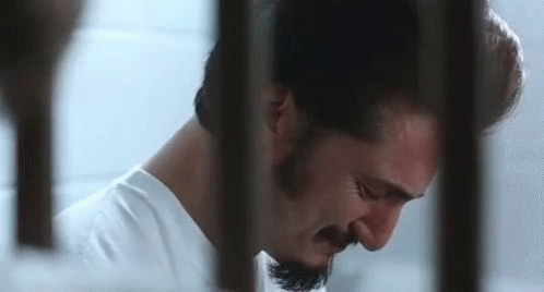 A Man With A Beard Is Behind Bars In A Jail Cell . GIF