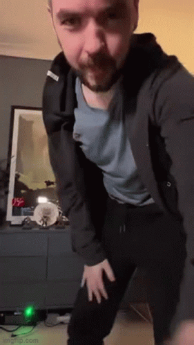 A Man With A Beard Is Dancing In A Living Room While Wearing A Black Jacket And A Blue Shirt . GIF