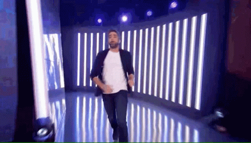 A Man With A Beard Is Dancing On A Stage With Striped Lights Behind Him . GIF