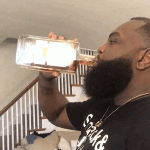 A Man With A Beard Is Drinking From A Bottle In Front Of A Staircase . GIF