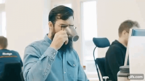 A Man With A Beard Is Drinking From A Cup In An Office . GIF