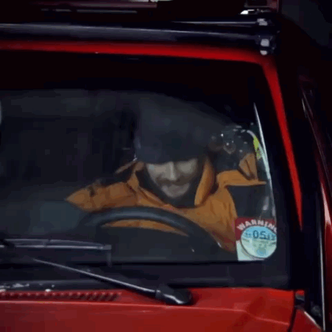 A Man With A Beard Is Driving A Red Car With The Words * Honk * Written On The Windshield GIF