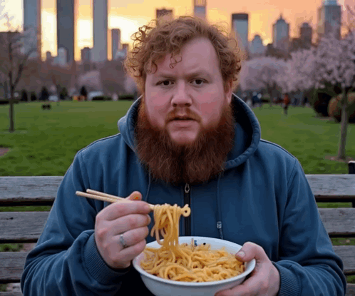 A Man With A Beard Is Eating Noodles With Chopsticks In A Park GIF