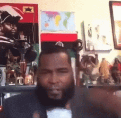 A Man With A Beard Is Giving A Thumbs Up In Front Of A Wall With A Map Of The World On It GIF