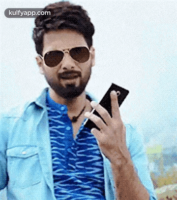 A Man With A Beard Is Holding A Cell Phone In His Hand . GIF