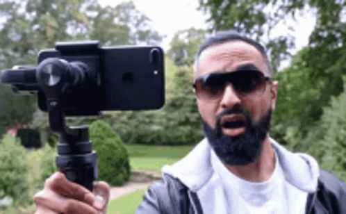 A Man With A Beard Is Holding A Cell Phone On A Tripod While Wearing Sunglasses . GIF