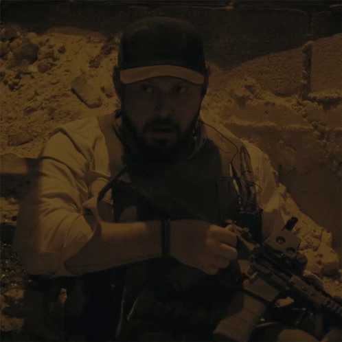 A Man With A Beard Is Holding A Gun And Wearing A Hat With The Letter T On It GIF