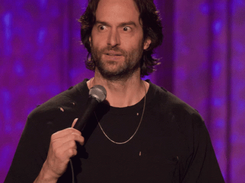 A Man With A Beard Is Holding A Microphone In Front Of A Purple Curtain GIF