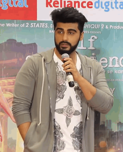 A Man With A Beard Is Holding A Microphone In Front Of A Sign That Says Reliance Digital GIF
