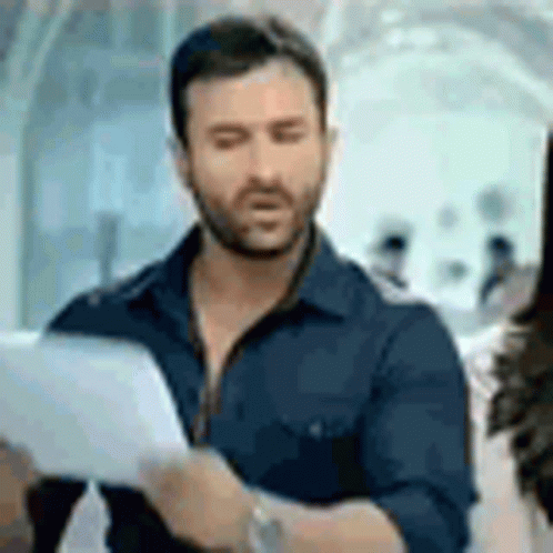 A Man With A Beard Is Holding A Piece Of Paper And Looking At It . GIF