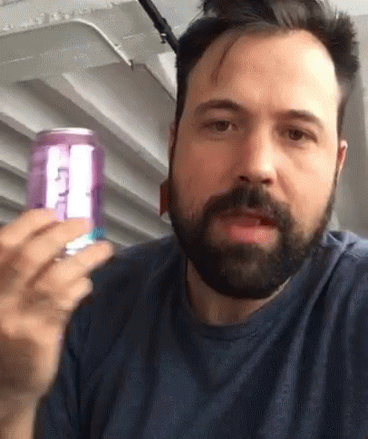 A Man With A Beard Is Holding A Purple Can Of Soda With The Letter T On It GIF
