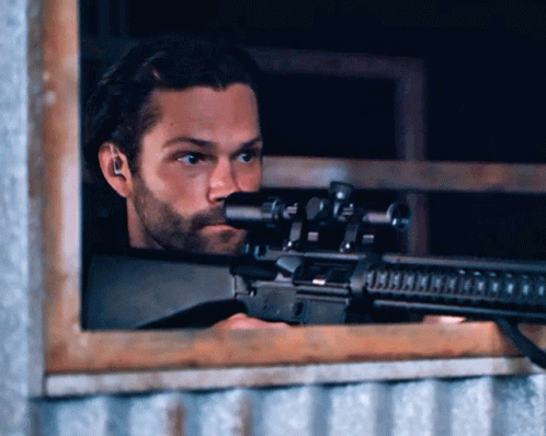 A Man With A Beard Is Holding A Rifle In Front Of A Window GIF